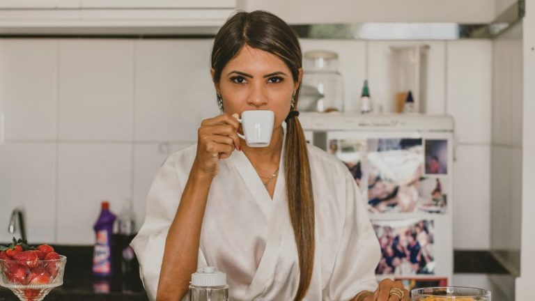 A woman in a white robe enjoys coffee with breakfast in a cozy kitchen setting.